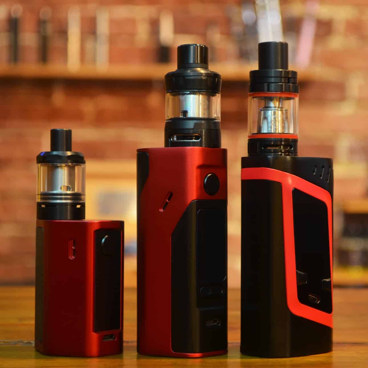 Vape devices with red and black design on a wooden surface against a brick wall background.